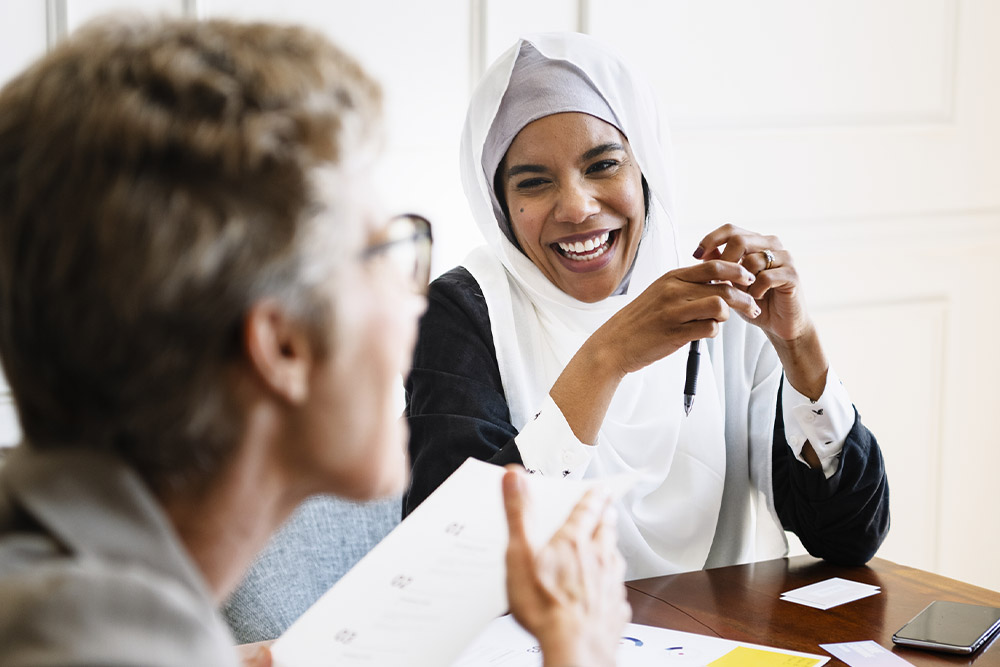 A woman in a white hijab smiles during a meeting, holding a pen. Papers and a phone are on the table.