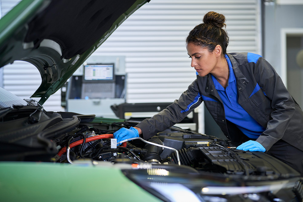  Technician in blue gloves working on the engine of a green Ford car with the hood open.