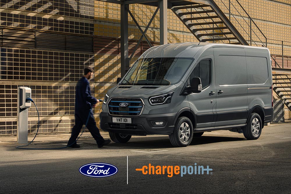 A person charges a grey Ford E-Transit van at an outdoor charging station.