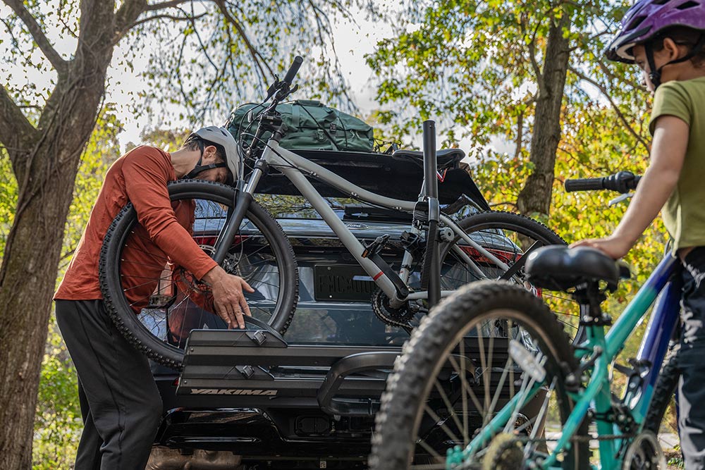 A person loads a bicycle onto a car's rear-mounted bike rack, with a child and another bicycle nearby.