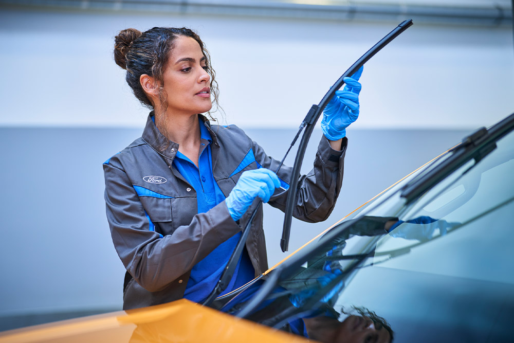 A Ford technician in blue gloves replaces a wiper blade on a car windshield in a service garage.