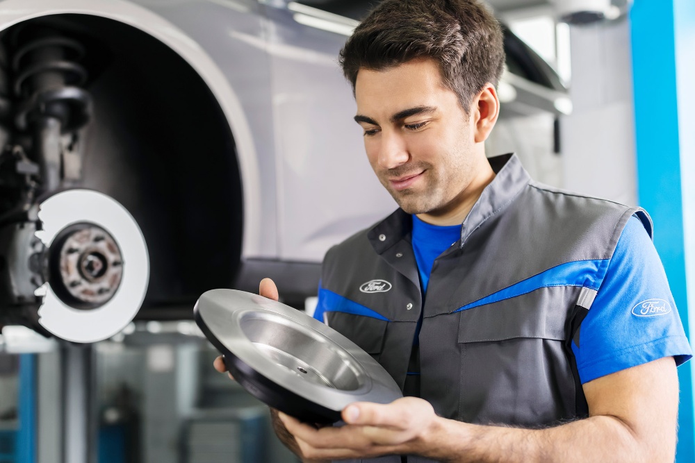 A technician in a Ford uniform inspects a brake disc in a service garage, with a car's wheel assembly in the background.