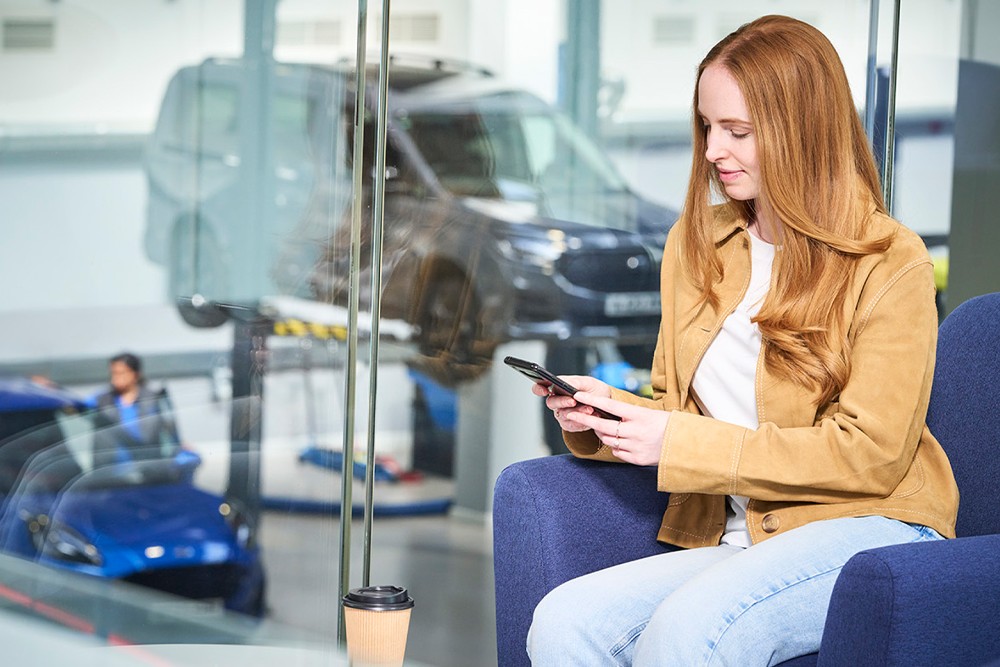 Person on phone in a Ford service waiting area, a van on a lift and a blue car are visible through the glass.