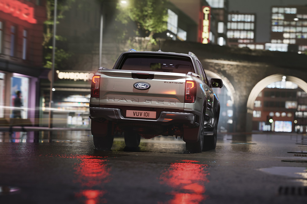 Back rear view of a silver Ford Ranger driving through a neon-lit cityscape at night.