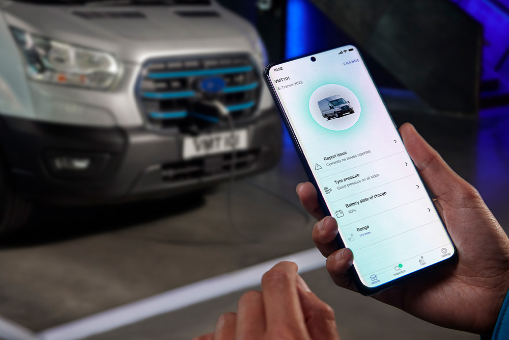A person holding a smartphone displaying an app interface, with a silver Ford Pro Transit van blurred in the background.
