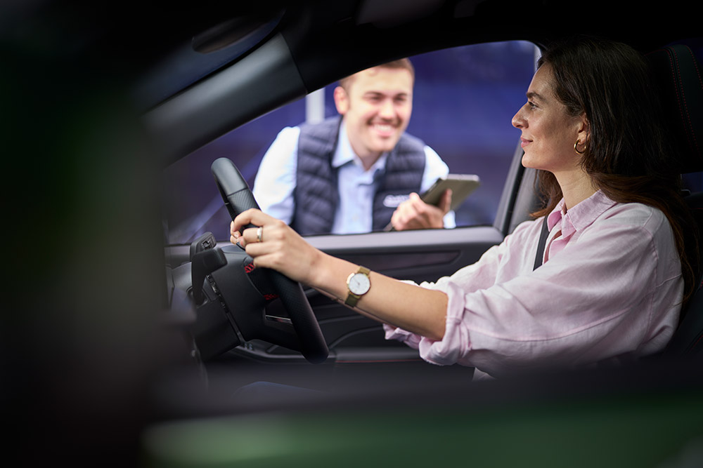  A person in the driver's seat of a Ford car, looking at a technician holding a tablet outside.