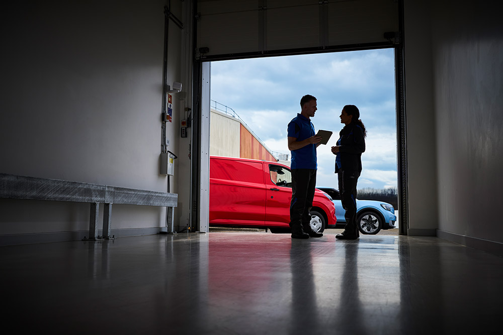 Two people talking in a garage doorway, with a red Ford Transit Custom and a blue Ford Capri outside.