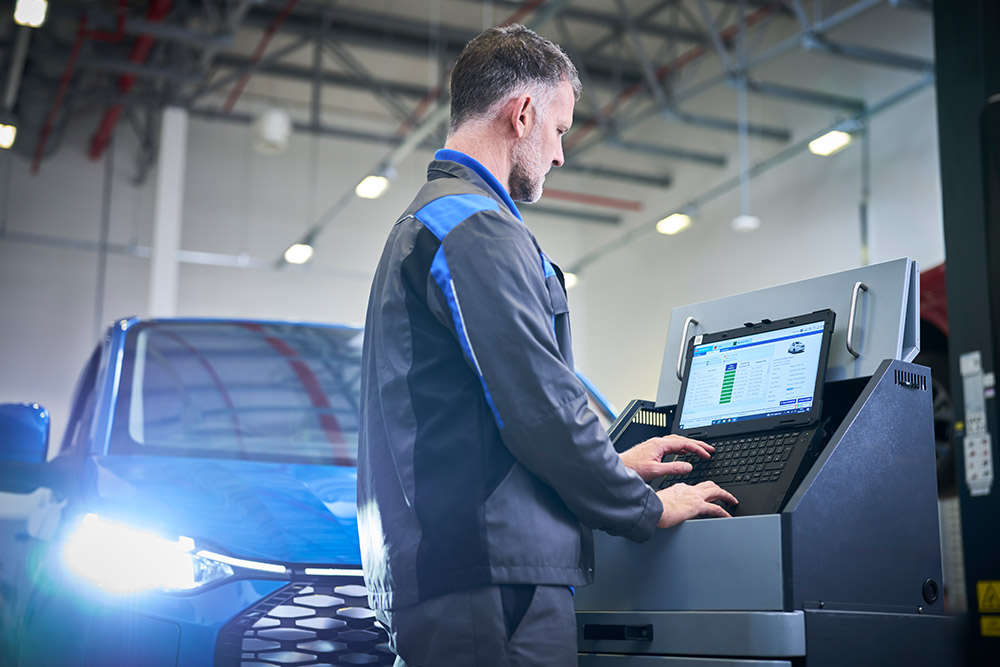A technician working on a laptop connected to a blue Ford Capri in a service bay.