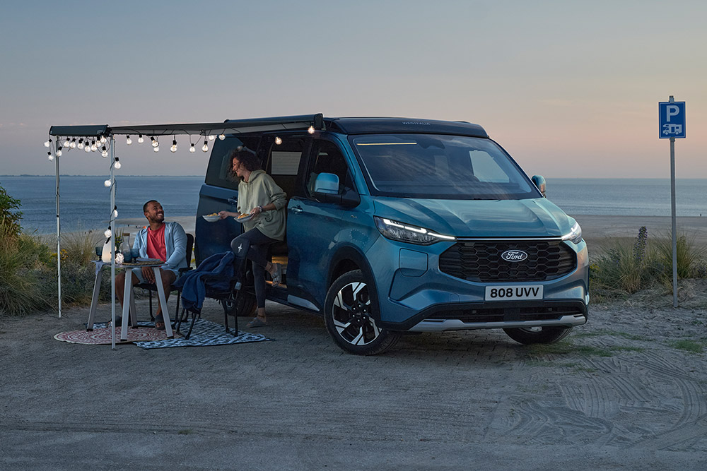 Two campers relaxing by a blue Ford Pro Transit Custom Nugget, a beachside parking area in the background.