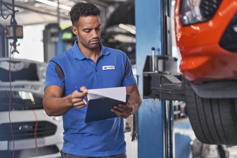 A Ford technician reviews documents on a clipboard in a service bay, with a car lifted on a hoist in the background.