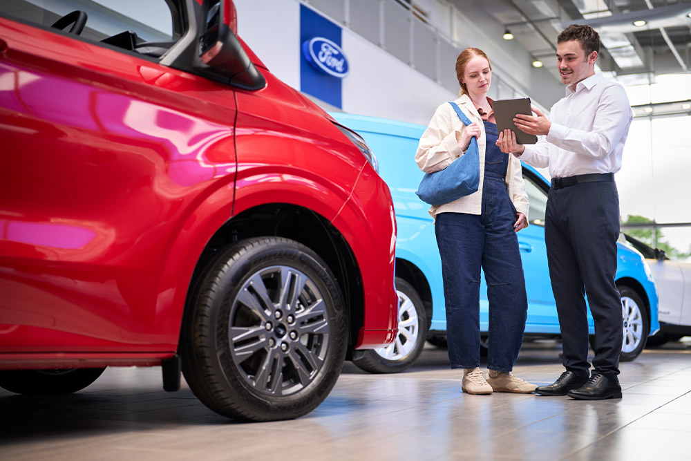 A Ford employee shows a tablet to a customer in a showroom with red and blue Ford vehicles.
