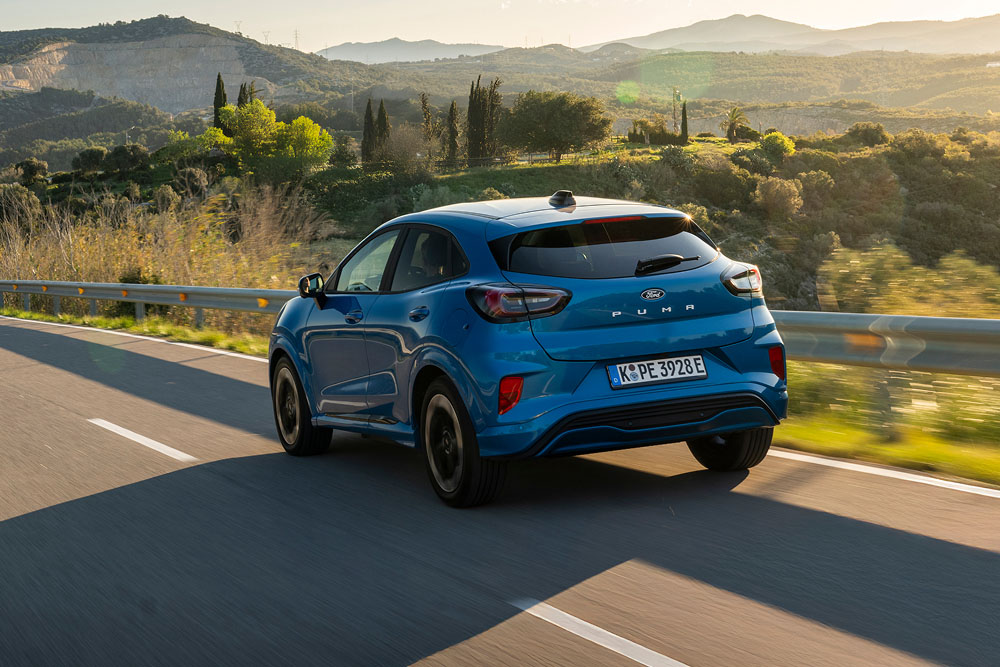 A blue Ford car driving on a winding road with mountains and trees in the background.