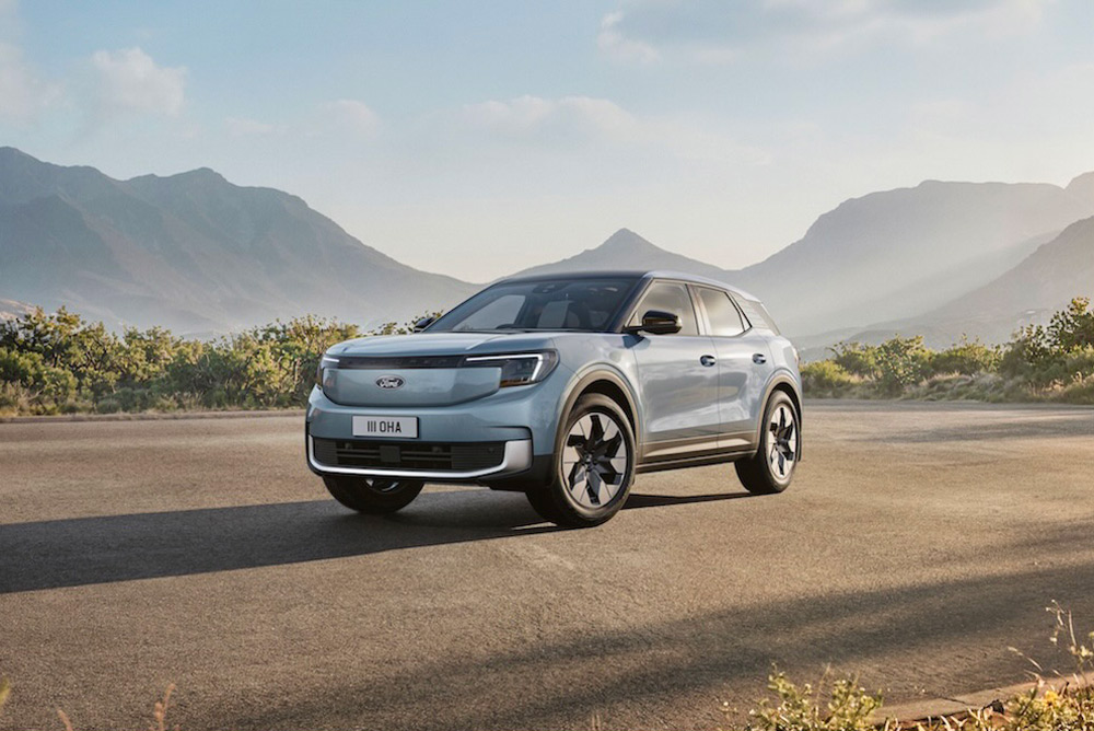 Front view of a light blue Ford Explorer EV parked on a paved road with mountains and trees.